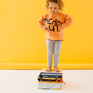 Baby Peanut Sweater by Castle. Child wearing Peanut Sweater Standing on Stack of Books Against Yellow Backdrop 